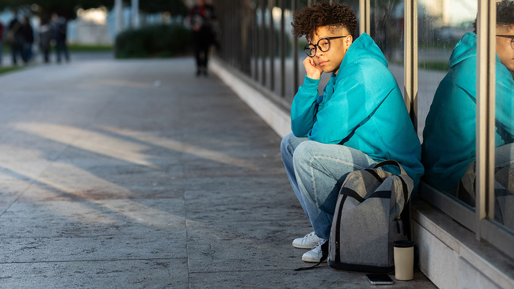 Jovem pensativo sentado sozinho com mochila, representando o impacto emocional e a importância da terapia online para brasileiros no exterior.